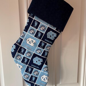 May include: A blue and light blue Christmas stocking with a repeating pattern of the University of North Carolina Tar Heels logo, the words "Carolina" and a footprint. The stocking has a dark blue cuff with a swirl pattern.