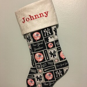 May include: A white Christmas stocking with a black and white New York Yankees pattern. The stocking has a white cuff with the name "Johnny" embroidered in red.