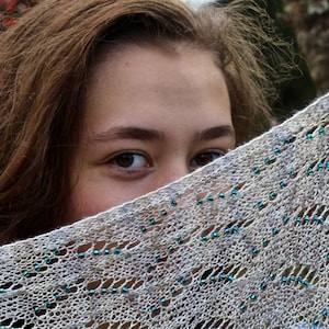 May include: A close-up of a woman's face partially obscured by a white crocheted shawl with blue beads. The shawl has a delicate, lacy pattern.