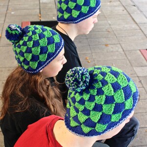 May include: Three people wearing blue and green checkered knit hats. The hats have a pom-pom on top.