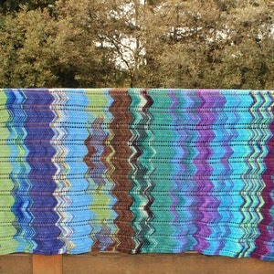 Op de afbeelding: Een gehaakt deken met een chevron-patroon in tinten blauw, groen, paars en bruin.