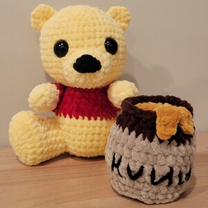 May include: A handmade crochet Winnie the Pooh plush toy with a matching honey pot. The bear is yellow with a red shirt, black eyes, and a black nose. The honey pot is gray with brown accents and the word "Hunny" in black.