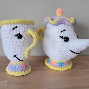 May include: Two handmade crochet figures: a teacup and a teapot. The teacup is white with a yellow rim and handle, and a colorful base. The teapot is white with a yellow handle, a purple and yellow lid, and a colorful base. Both have black eyes and smiling faces.