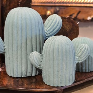 May include: Three light blue cactus sculptures of varying sizes. Each cactus has a textured surface and ribbed detailing, with curved arms extending from the main body. The sculptures are arranged on a dark brown surface, creating a desert-inspired decorative theme.