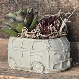 May include: A gray concrete planter shaped like a vintage Volkswagen bus, filled with green and purple succulents.