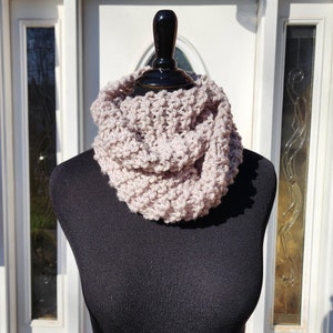 May include: A light pink knitted infinity scarf on a black mannequin. The scarf is made of a soft, chunky yarn and has a simple, elegant design.