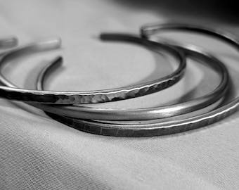 May include: Three silver bracelets stacked on top of each other. The bracelets are all different widths and have a hammered texture.