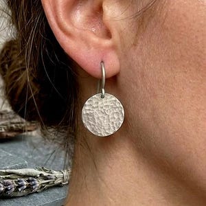 May include: A silver-toned earring featuring a circular pendant with a textured, hammered finish. The round disc hangs from a simple metal hook, creating a drop-style design that catches the light across its uneven, reflective surface.