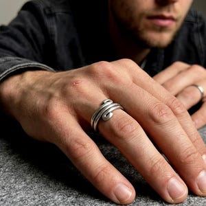 Op de afbeelding: Een zilverkleurige ring met een bypass-ontwerp met twee overlappende banden die om de vinger wikkelen. Het metaal heeft een gladde, gepolijste afwerking met een afgerond profiel, wat een continu spiraaleffect op de vinger creëert.