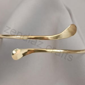 May include: A gold-toned, open-ended bangle bracelet with a simple, curved design. The bracelet is made of two thin, smooth bands that wrap around the wrist.