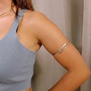 Può includere: Un bracciale da braccio in metallo color argento con un design semplice a fascia aperta e punte arrotondate. Il bracciale è indossato sulla parte superiore del braccio, abbinato a una canotta a coste azzurra e una sottile collana color oro.