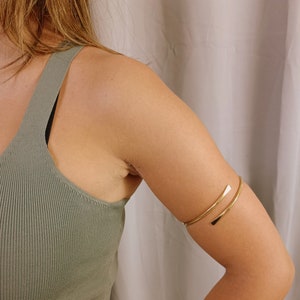 May include: A gold-toned metal arm cuff bracelet. The bracelet is open and has a simple, minimalist design.