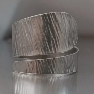 Puede incluir: Un anillo envolvente plateado con una superficie texturizada. El anillo tiene una banda ancha que se envuelve alrededor del dedo, creando un diseño moderno y único. La superficie texturizada añade interés visual.
