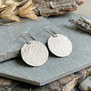 May include: A pair of silver-toned circular earrings featuring a hammered, textured surface. Each earring is suspended from a simple curved wire hook, creating a dangling design that catches the light across the uneven, metallic finish.
