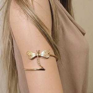 Op de afbeelding: Een goudkleurige metalen armband voor de bovenarm met een gedetailleerd libelle-ontwerp. De libelle heeft gestructureerde vleugels en een slank lichaam dat naar beneden loopt en aansluit op de gladde, gebogen band die om de bovenarm sluit.