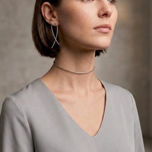 Peut inclure: Un collier ras-du-cou en métal argenté composé d'une fine bande circulaire lisse qui repose près du cou, accompagné de boucles d'oreilles assorties en métal argenté présentant un design fin et incurvé qui se croise sous le lobe de l'oreille.