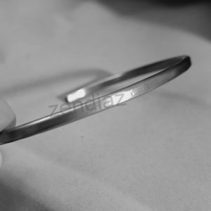 Könnte beinhalten: Ein silberfarbenes, offenes Armband. Das Armband hat eine glatte, polierte Oberfläche und ein schlichtes, minimalistisches Design. Das Wort "zendiaz" ist auf dem Armband zu sehen. Das Armband wird vor einem unscharfen Hintergrund gezeigt.