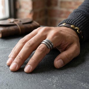 May include: A silver-toned metal ring featuring a wrap-around design with a series of vertical notches along the outer edges. The ring has a brushed, matte finish and is worn on the middle finger, showcasing a layered, industrial-inspired aesthetic.