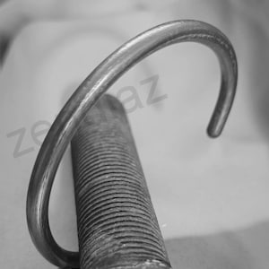 Może przedstawiać: Srebrzysta metalowa bransoletka o zakrzywionym, otwartym designie. Bransoletka jest pokazana w zbliżeniu, podkreślając jej gładką, wypolerowaną powierzchnię. Bransoletka spoczywa na teksturowanym, cylindrycznym obiekcie.