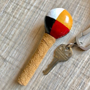 May include: A hand-painted maraca with a tan leather handle and a colorful spherical head. The head is divided into white, yellow, red, and black sections. A silver key and keychain are also in the image.