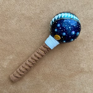May include: A hand-painted maraca. The dark blue sphere is decorated with white dots and blue orbs, resembling a starry night. The top has a geometric pattern in white and teal. The handle is wrapped in tan leather.