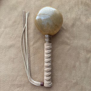 Pode incluir: Uma maraca artesanal com uma cabeça esférica castanha clara e uma pega enrolada. A pega é enrolada num padrão em ziguezague com uma tira de couro e uma pequena conta de madeira. Um feixe de tiras de couro pende da base.