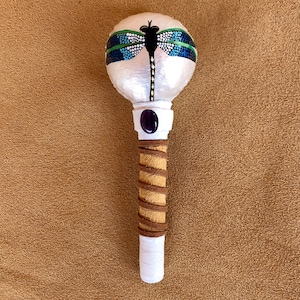 May include: A handcrafted rattle featuring a white, spherical head adorned with a black dragonfly design and blue and green accents. The handle is wrapped in tan and brown fabric, with a white base and a purple gem.