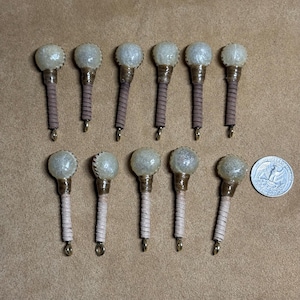 Puede incluir: Una colección de objetos decorativos en miniatura con forma de maza. Cada uno tiene una cabeza esférica texturizada y un mango envuelto, con un pequeño lazo de metal en la base. La paleta de colores incluye tonos beige, marrón y plateado, con una moneda como referencia de tamaño.