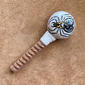 May include: A white rattle with a black and white spider design. The rattle has a brown leather handle.