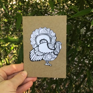 May include: A black and white line drawing of a turkey with intricate patterns on its feathers. The turkey is facing right and has its head turned to the side. The drawing is on a brown card.