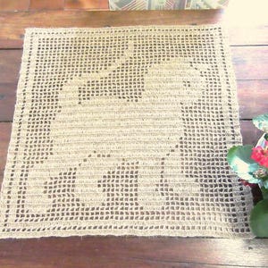 May include: A square, beige crochet doily featuring a grid-like pattern with a central elephant silhouette formed by denser stitching. The piece has a flat, open-work design with a solid border frame, showcasing a natural fiber texture throughout.
