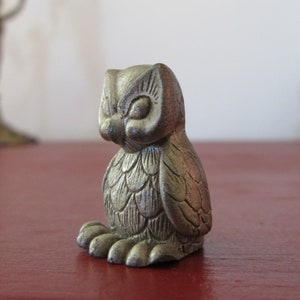 Puede incluir: Pequeña figura de búho metálico con detalles de plumas y ojos. El búho está posado sobre una superficie roja oscura. La figura tiene una apariencia vintage o antigua.