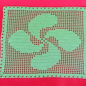 May include: A handmade, square, light green crochet doily with a central design. The design features a stylized, symmetrical pattern. The doily is set against a solid red background, creating a striking contrast. The crochet work is detailed and open.