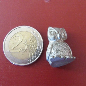 Peut inclure: Une petite figurine de hibou argentée avec des détails de plumes et de visage. Le hibou est à côté d'une pièce de 2 euros pour la comparaison de taille. La figurine est sur une surface rouge.