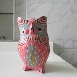 May include: A hand-painted wooden owl figurine featuring a vibrant pink base color with intricate blue and black feather detailing. The owl has large, expressive eyes, a pointed beak, and a yellow accent mark on its chest, standing upright.