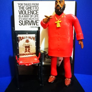 Op de afbeelding: Een rood en zwart action figure van een man die een rood shirt, een rode broek en een gouden ketting met een hanger draagt. De figuur staat op een zwarte basis met een zwarte en rode stoel en een bord met de tekst "Death Row Records". Het bord bevat ook de quote "For thugs from the ghetto, violence is a way of life. It's what helps you survive" - Suge Knight.