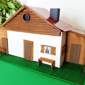 Può includere: Modello di casa in miniatura con tetto marrone, camino e porte in legno. La casa ha una facciata bianca e poggia su una base verde, con una piccola panchina in legno. Una piccola recinzione è sul lato.