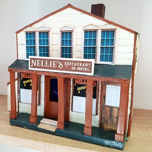 May include: A detailed miniature model of Nellie's Restaurant Hotel. The building features a cream-coloured exterior with brown trim and a dark green roof. The sign above the porch reads "Nellie's Restaurant Hotel". The model has three windows on the second floor.