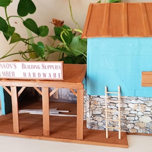 Può includere: Un modello in miniatura di un negozio di materiali edili con facciata blu, tetto marrone e fondamenta in pietra. L'insegna recita "Hanson's Building Supplies Lumber Hardware". Una piccola scala di legno è appoggiata all'edificio.