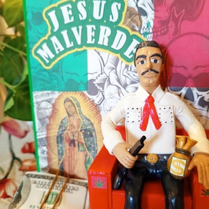 Può includere: Una statuetta di un uomo seduto su una sedia rossa, che tiene una pistola e una borsa di denaro. La statuetta indossa una camicia bianca con accenti rossi, pantaloni neri e una bandana rossa. Lo sfondo presenta una bandiera messicana verde e rossa con le parole "JESUS MALVERDE" in lettere gialle.