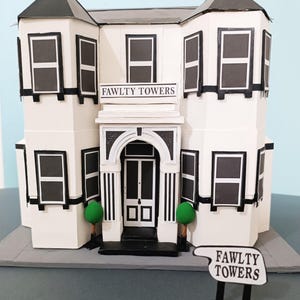 Può includere: Un modello di carta bianco di Fawlty Towers con finiture nere per finestre, tetto e bordi. L'edificio presenta un'insegna con la scritta "FAWLTY TOWERS" e un'altra insegna separata con lo stesso testo.