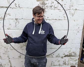 Op de afbeelding: Een grote, dunne, cirkelvormige metalen hoepel met een donkergrijze afwerking. De hoepel is gemaakt van één doorlopend stuk metaal en vormt een perfecte ring die rechtop wordt gehouden, wat het eenvoudige industriële ontwerp benadrukt.