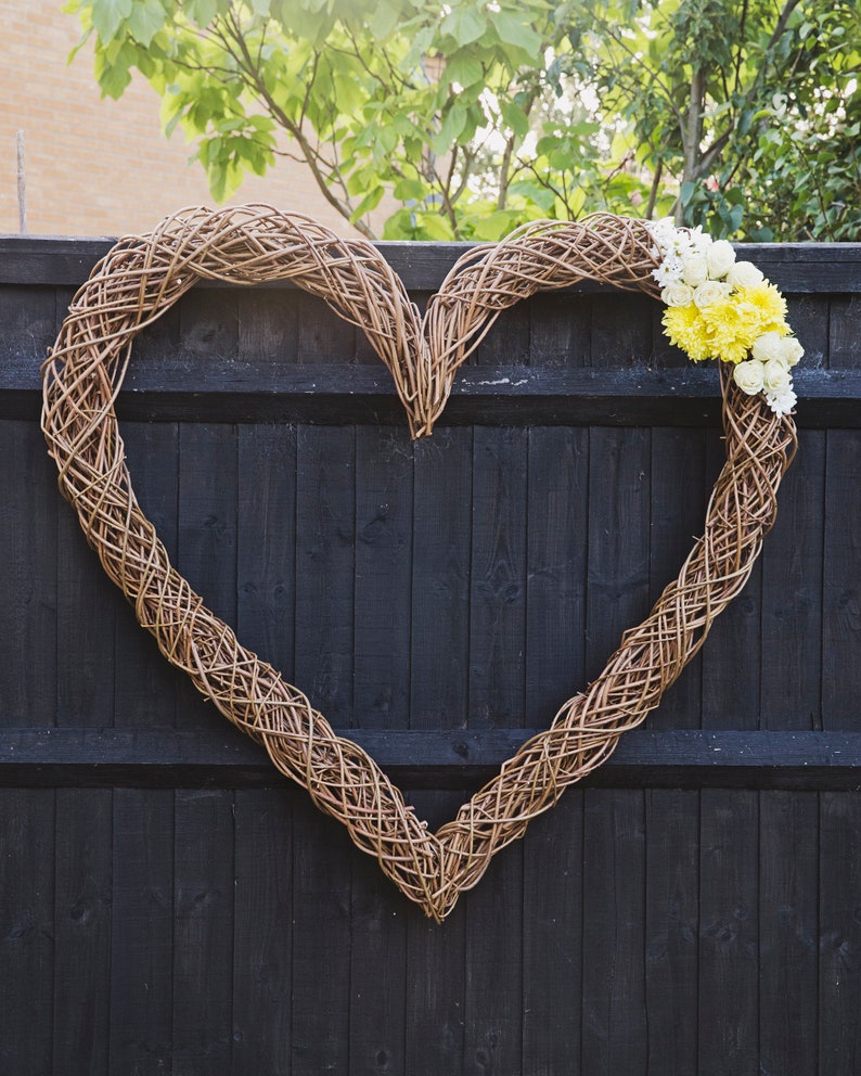 Op de afbeelding: Een geweven wilgenhartenkrans versierd met witte en gele bloemen, hangend aan een zwart houten hek.