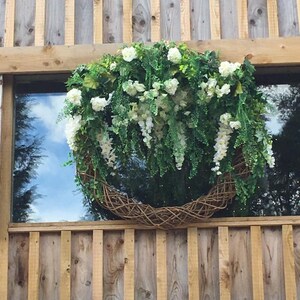 Op de afbeelding: Een grote, ronde krans gemaakt van gevlochten takken en groen, versierd met witte bloemen en overhangend groen. De krans hangt aan een houten muur.