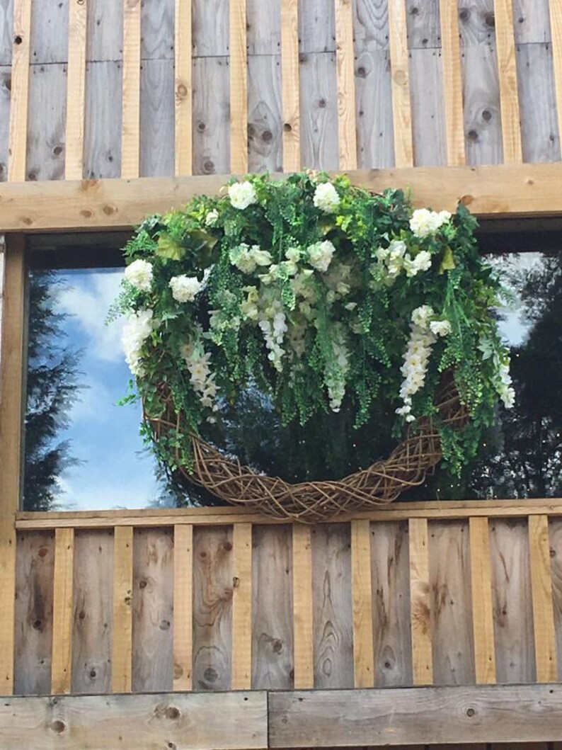 Op de afbeelding: Een grote, ronde krans gemaakt van gevlochten takken en groen, versierd met witte bloemen en overhangend groen. De krans hangt aan een houten muur.
