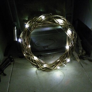Op de afbeelding: Een bruine wilgenkrans met witte LED-lampjes eromheen gewikkeld. De krans staat voor een zwarte open haard.