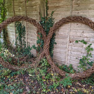 Op de afbeelding: Twee grote, geweven wilgenkransen, elk met een ronde vorm, leunen tegen een houten hek. De kransen zijn gemaakt van natuurlijke bruine wilgentenen en zijn met elkaar verweven om een decoratief ontwerp te cre&euml;ren.