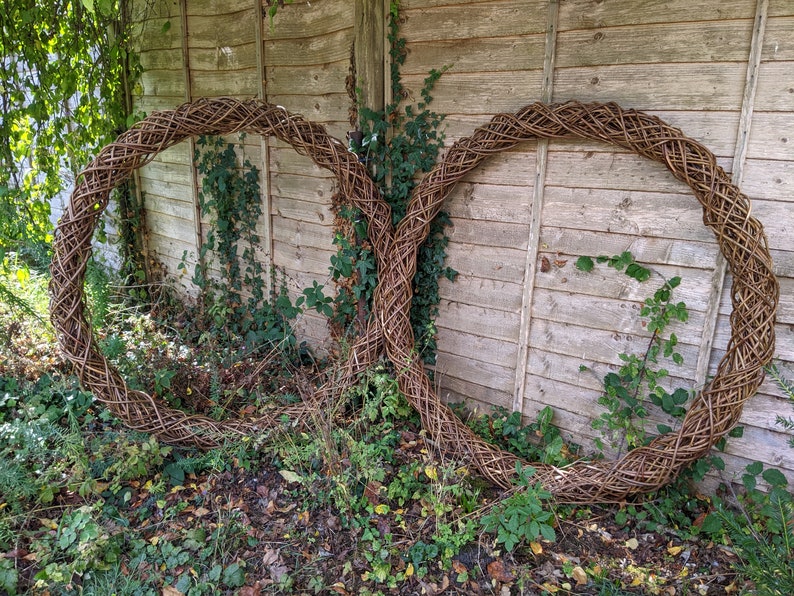 Op de afbeelding: Twee grote, geweven wilgenkransen, elk met een ronde vorm, leunen tegen een houten hek. De kransen zijn gemaakt van natuurlijke bruine wilgentenen en zijn met elkaar verweven om een decoratief ontwerp te cre&euml;ren.