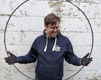 Op de afbeelding: Een grote, dunne, cirkelvormige metalen hoepel met een donkere, matte afwerking. De hoepel wordt opengehouden door twee handen met zwarte werkhandschoenen, wat het eenvoudige, doorlopende en onversierde ontwerp benadrukt.