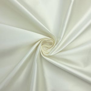 Peut inclure: Un tissu blanc cassé uni avec une finition lisse et lustrée. Le matériau est rassemblé au centre, créant des plis doux et rayonnants ainsi que des ombres profondes qui soulignent la texture réfléchissante et satinée de la surface textile.